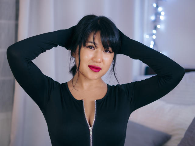 ErikaKim has black hair, red lipstick, and wears a black long-sleeve zip-up top, hands in her hair, smiling at the camera. Her tits are small, her pussy is unseen.