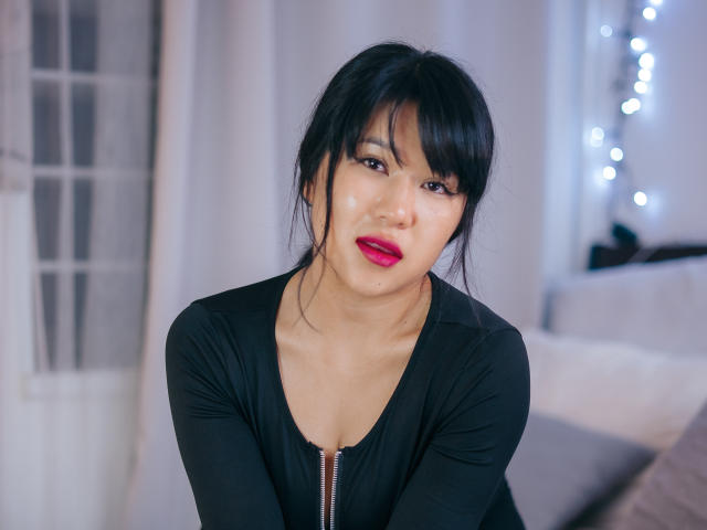 ErikaKim, con cabello negro, lápiz labial rojo y una blusa negra con una cremallera, sentada y sonriendo suavemente. Sus glúteos no son visibles. Tiene un rostro lindo y atractivo..