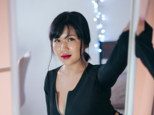 ErikaKim with black hair, red lipstick, and a black zip-up shirt, standing in front of a mirror, holding her shirt. Her tits are small and she's looking cute.