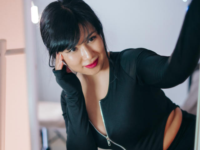 ErikaKim with black hair, bangs, and red lipstick, wearing a tight black zip-up top, showing her tits through cut-out sides, leaning forward with a sexy smile.