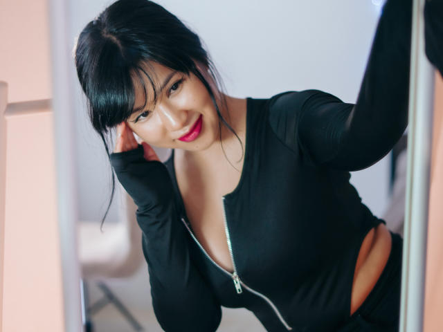 ErikaKim met zwart haar, rode lippenstift en een zwart top met rits, leunt tegen een muur, glimlachend, toont haar borsten, hand in haar haar, sexy, aantrekkelijk, medium huidskleur, schattig..