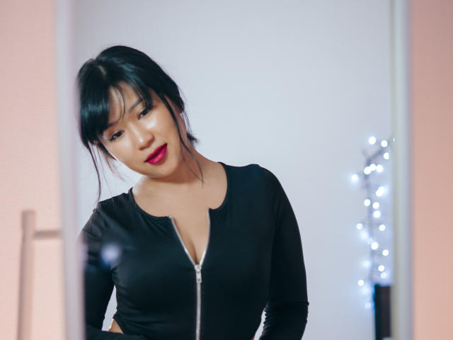 ErikaKim met zwart haar, rode lippenstift en een zwart topje met rits, dat haar decolleté laat zien, staat iets links, met een subtiele glimlach. Vage voorgrond..