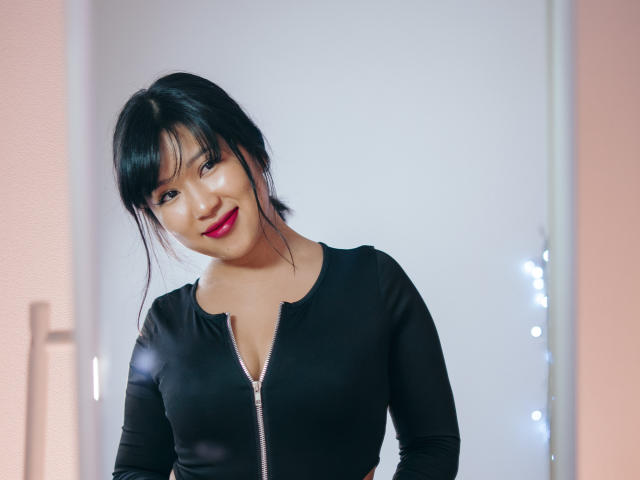 ErikaKim, avec ses cheveux noirs, son rouge à lèvres rouge et son haut noir zippé moulant, sourit à l'appareil photo, montrant sa petite poitrine et un visage mignon et sexy..