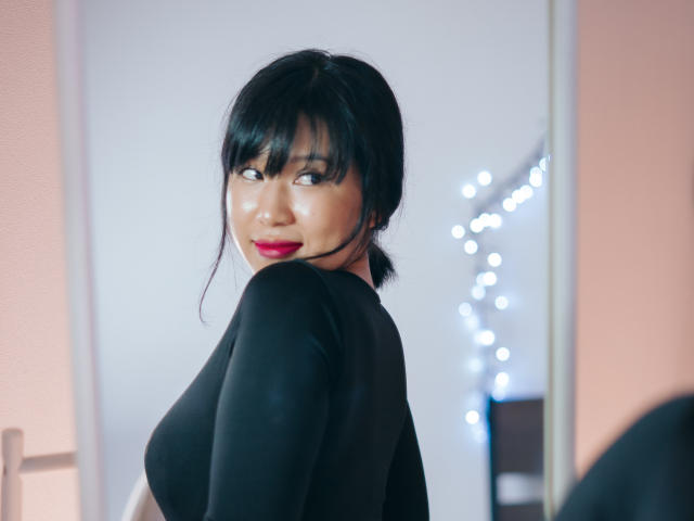 ErikaKim met zwart haar in een staart, gekleed in een zwarte top met lange mouwen, kijkt over haar schouder, rode lipstick, lachend, spiegelbeeld, zachte bokeh-lichtjes op de achtergrond..