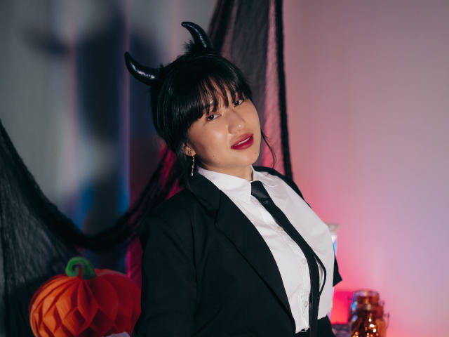 ErikaKim, avec des cornes noires, du rouge à lèvres, et un costume noir, montrant ses seins, a de longs cheveux noirs, et une citrouille sur la gauche, dans une pièce sombre et éclairée..