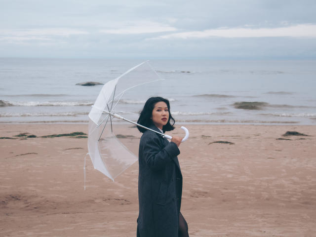 ErikaKim, avec des cheveux noirs, portant un manteau sombre, tenant un parapluie transparent, debout sur une plage de sable, regardant sur le côté, avec un océan calme et nuageux en arrière-plan..