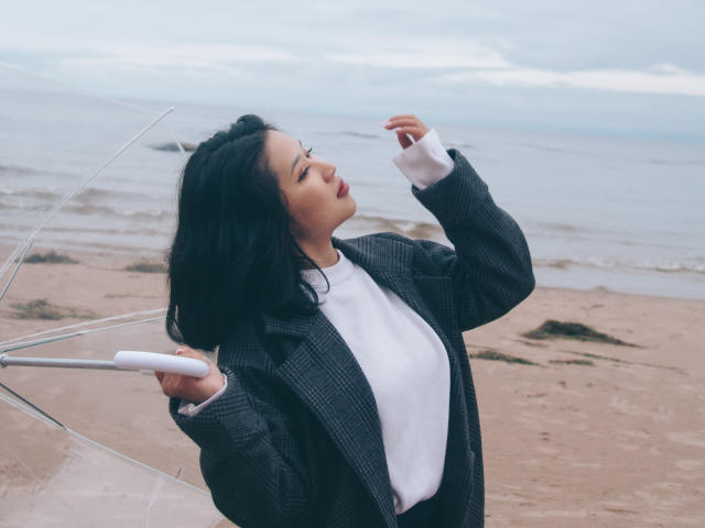 ErikaKim with black hair, wearing a plaid jacket and white shirt, holding a clear umbrella, looking up, her hand near her mouth, standing on a beach.