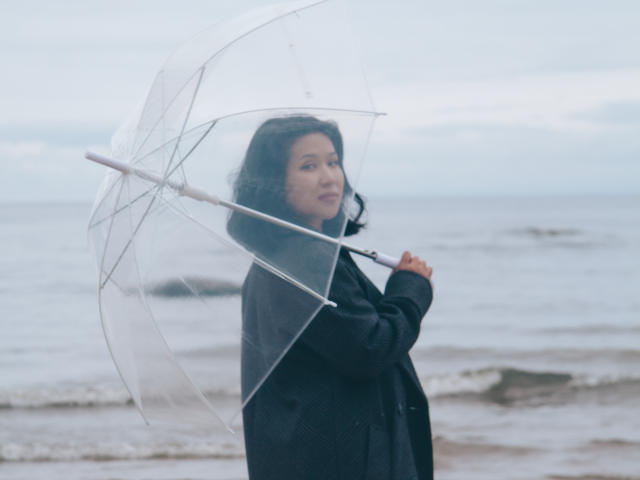 ErikaKim, avec ses cheveux noirs bouclés, tenant un parapluie transparent, portant un manteau noir, debout sur une plage mouillée, regardant de côté avec un léger sourire..