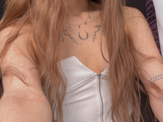 Karish's chest with long brown hair, white zip-up dress, crescent moon tattoo, necklace, and small tattoos on her right arm. Boobs visible, no other body parts shown.
