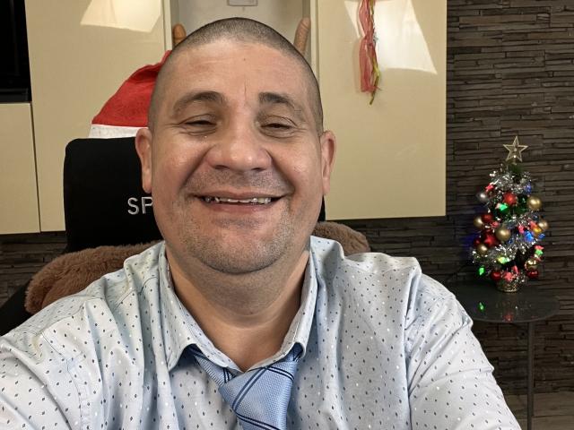 CrisSalon, a bald, middle-aged man with a rounded face and light skin, smiles broadly. He wears a light blue, patterned shirt with a loose blue tie. His short-cropped hair is completely shaved. A small, colorful Christmas tree with a star topper sits on a glass table to his right. A brown fuzzy chair is partially visible behind him. The background includes a beige wall and a textured dark brown stone-like wall section. The photo is a casual selfie.