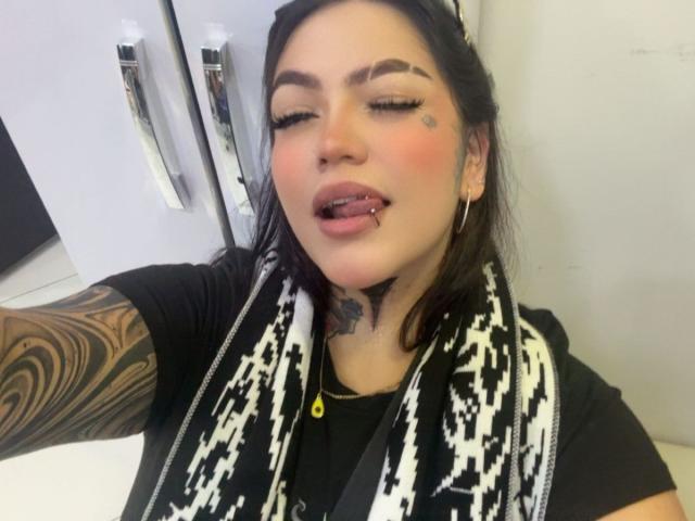 TessaMagic with black hair, tattooed arm, black shirt, white/black scarf, tongue piercing, and hoop earrings, looking seductive with closed eyes and slightly open mouth.