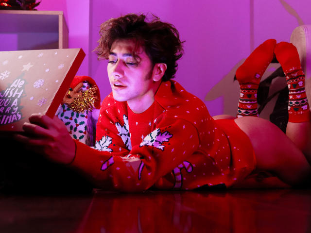 TonyDan allongé par terre, portant un pull de Noël rouge, des chaussettes rouges, sans pantalon, regardant un cadeau, éclairage violet, cheveux courts et en désordre, maquillage bleu pour les yeux, rouge à lèvres rouge, expression de surprise..