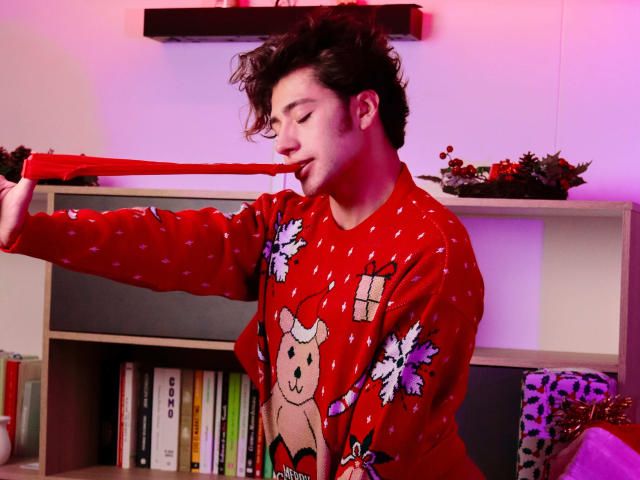 TonyDan, avec ses cheveux bruns bouclés, portant un pull de Noël rouge, tenant un ruban rouge dans sa bouche, regardant vers le bas avec un léger sourire, se tenant dans une pièce confortable..