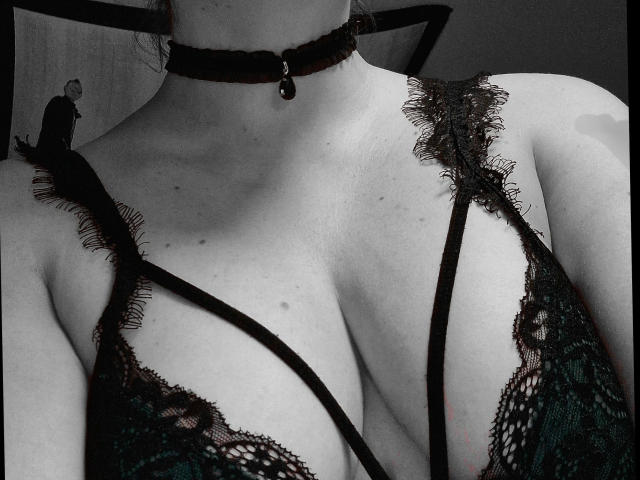 Curvyhoneyx-hot wearing a black lace bra with deep cleavage, black choker with a dangling bell, and tiny man figurine on her shoulder strap. Monochrome, sexy, detailed.