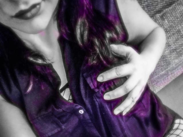 Curvyhoneyx-hot with dark purple, shiny, silky top, hand on chest, dark lipstick, wavy black and purple hair, partial view of face and chest, monochrome background.