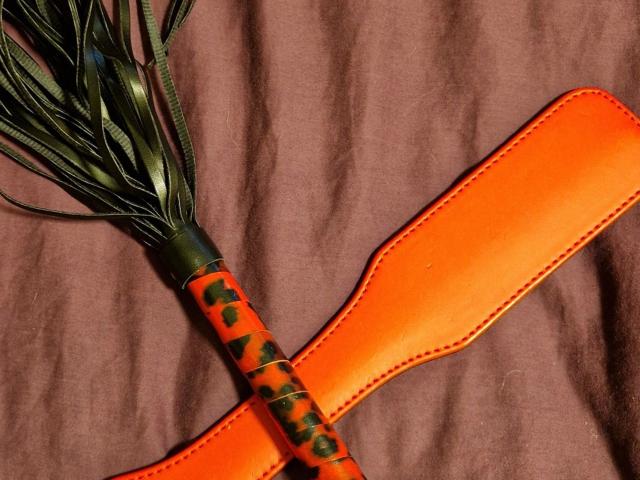 A bright orange flogger with black patterned handle and green leather tails is laid on a brown fabric. Curvyhoneyx-hot's toy.