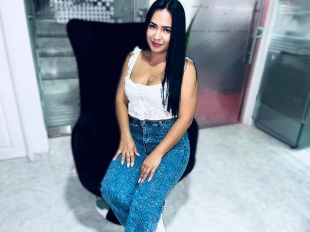 HannaRoux with long black hair, white lace top, showing her tits, and blue jeans, sitting on a black chair, smiling, hands on her knees, in a bright room.