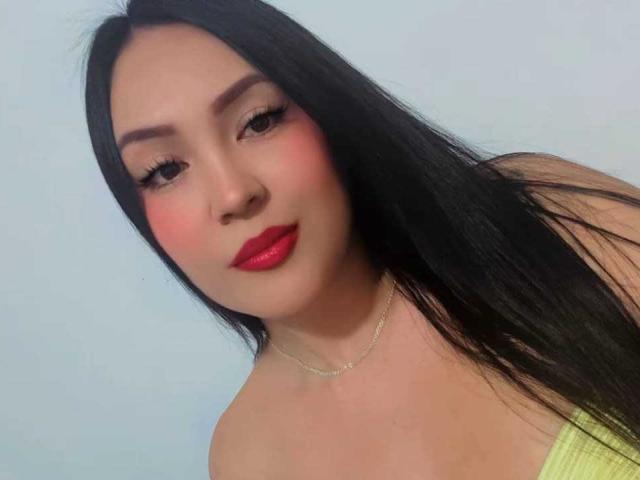HannaRoux with long black hair, red lipstick, pink blush, and a green strap top, showing her tits and a necklace against a plain background.
