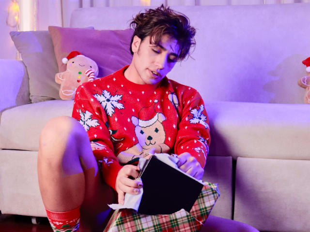 TonyDan, con cabello castaño corto, vistiendo un suéter navideño rojo con un osito de peluche y copos de nieve, sentado en un sofá, mirando un libro negro, falda escocesa, calcetines rojos..