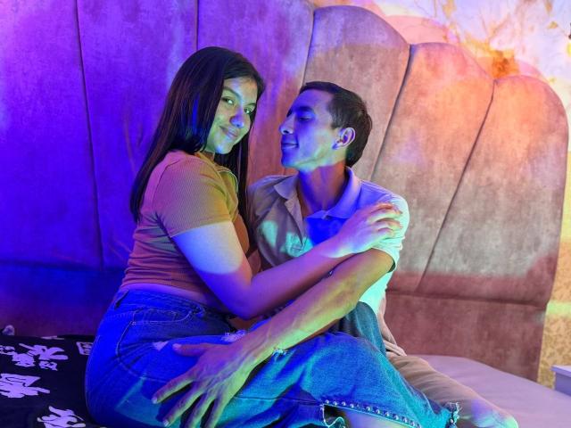 AdanNdEva with long black hair sits on a bed, facing a man with short dark hair. She's wearing a brown t-shirt and blue jeans, her hands on his chest. He's in a beige polo shirt and blue jeans, looking at her with a slight smile. Blue and purple lighting casts a cool glow. The bed has a floral-patterned black blanket. AdanNdEva's right hand rests on his thigh. The man's right hand is on her back.