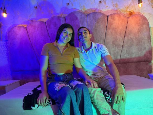 AdanNdEva, a woman with dark hair in a yellow crop top and blue jeans, and AdanNdEva, a man with short hair in a white polo shirt and beige pants, sit on a bed with a patterned black blanket. They're under blue and purple lights, with a large, curved, beige headboard behind them. The man's left hand rests on her right knee. The man's right hand is on the bed. The woman's expression is relaxed, the man's is slightly curious. Image has a "BRAZZERS.COM" watermark.