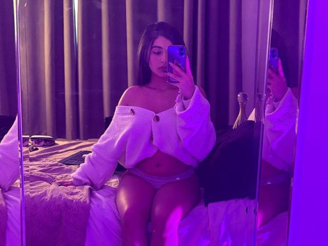 EmmaBeaumont takes a mirror selfie in a dimly lit room with purple lighting. She sits on a bed, wearing an off-shoulder white sweater and pink lace panties. Her long dark hair is straight, and she holds a blue phone in her right hand. Her left hand rests on the bed. Reflections in the mirror show her from the side. The bed has a brown and white striped blanket, and a black pillow is visible. A silver candlestick is on the right. Her expression is neutral.