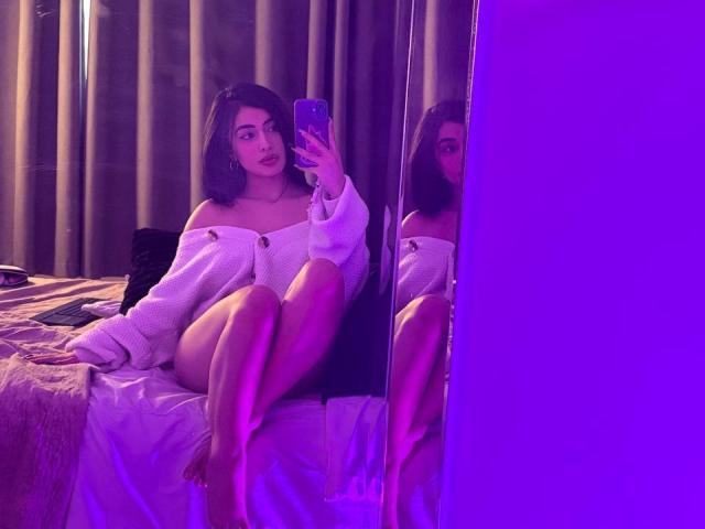 Emma Beaumont, com seios médios, sentada na cama, vestindo um suéter branco com um ombro descoberto, segurando um telefone, refletida no espelho, iluminação roxa no quarto, cabelo ondulado escuro, pernas cruzadas, olhando para a câmera..
