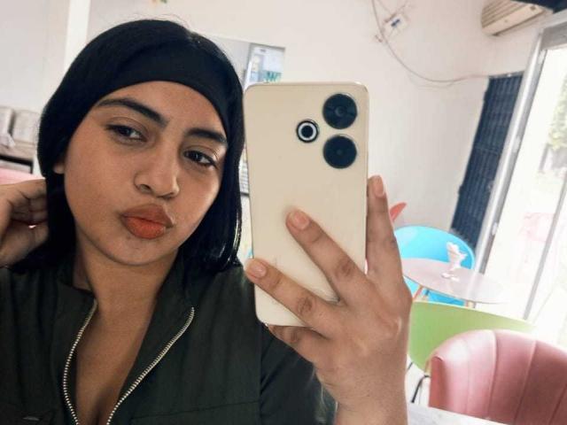 SofiaAngels takes a selfie with a white iPhone, holding it in her right hand. She has long black hair, a black headband, and wears a green jacket with a zipper. Her lips are puckered in a small kiss. She has a nose ring and neatly groomed eyebrows. The background includes a pink chair, a blue and green table, and a window with daylight. The image is clear and focuses on SofiaAngels' face and the iPhone.