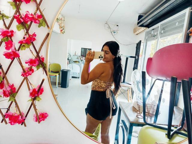 Sofia: Een vrouw met donker haar, een gebruinde huid en een rond gevormd achterwerk, gekleed in een kanttop en zwarte shorts, die een spiegelzelfportret maakt in een lichte kamer met roze bloemen..