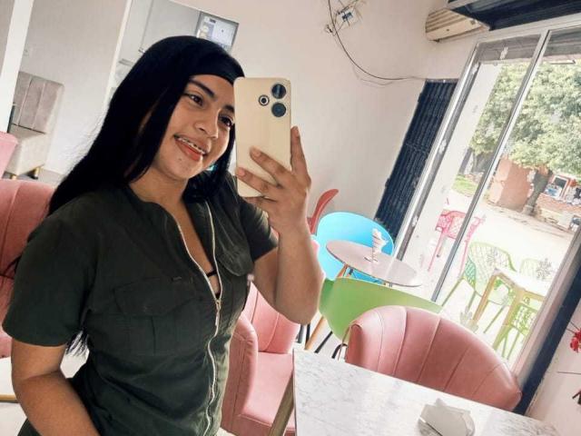 Sofia. Ángeles con cabello negro largo, sonriendo, sosteniendo un teléfono, vistiendo una camisa verde, mostrando ligeramente sus senos, en una cafetería con sillas rosas y una puerta de vidrio..