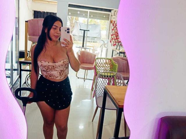 Sofia, une jeune femme aux longs cheveux noirs, portant un haut moulant à fleurs roses et une jupe noire à pois, tenant un téléphone pour un selfie devant un miroir, montrant sa poitrine généreuse et ses fesses..