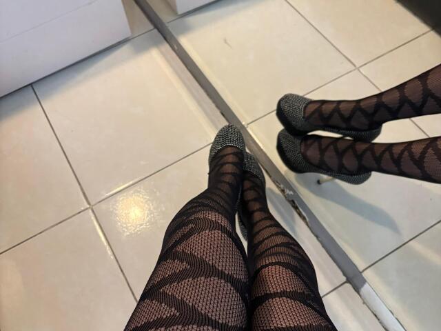 AnyaJay's legs in black fishnet stockings with diamond patterns, wearing sparkly silver heels, standing on white tiled floor. Her reflection is visible in the lower right.