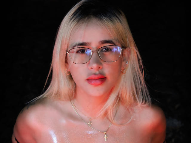 AndreaBoob with blonde hair, glasses, and a cross necklace, has shiny skin and is topless, showing her bare shoulders and slightly parted red lips in a dark setting.
