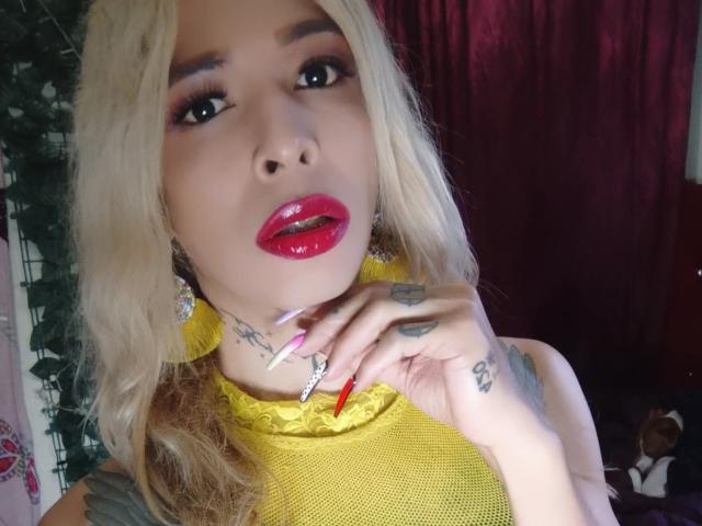 FlaquitaRaquel with blonde hair, red lips, yellow mesh top, and long red nails, showing tattoos on fingers and neck, looking up with a slight open-mouth expression.