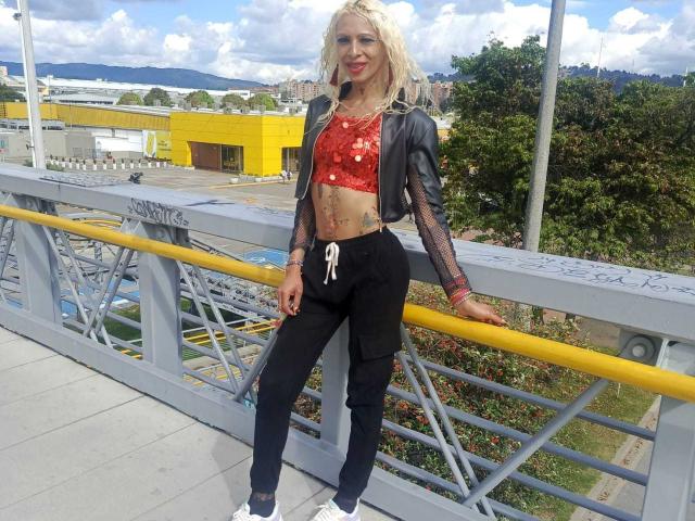 FlaquitaRaquel stands on a grey metal bridge, wearing a black leather jacket over a red floral crop top, black jogger pants, and white sneakers. She has blonde curly hair, red lipstick, and tattoos on her left side. She leans on a yellow rail with her right hand, smiling. The background includes greenery, a yellow building, and a cloudy sky. Her outfit contrasts with the grey bridge and yellow rail.