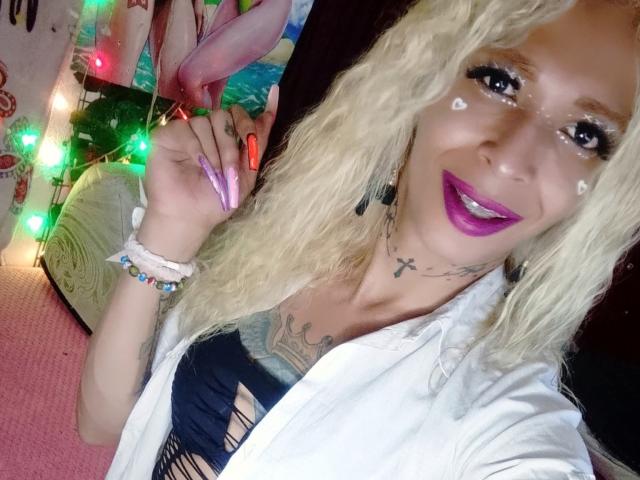 FlaquitaRaquel with blonde curly hair, purple lipstick, and white face paint dots, holding a pink dildo, wearing a white shirt, and blue ripped top, showing tattoos and long nails.
