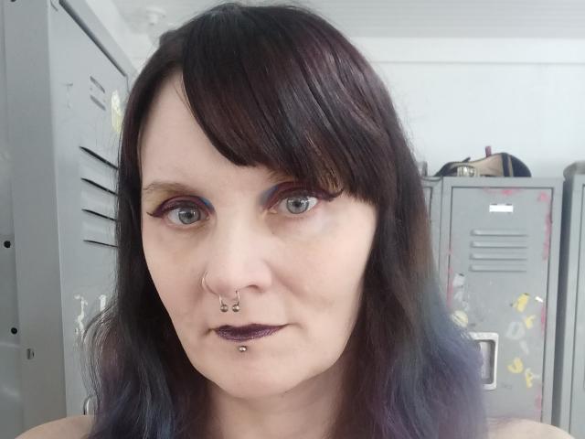 HotModellX with pale skin, dark brown wavy hair, blue eyeshadow, black eyeliner, purple lipstick, nose ring, and lower lip piercing, standing in front of gray metal lockers.
