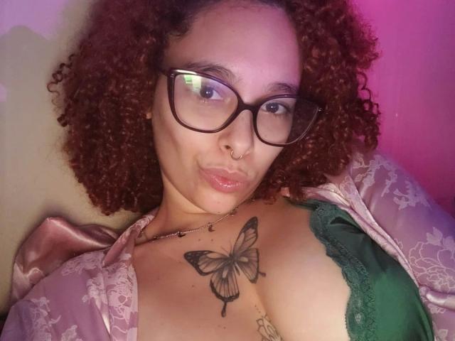 LizzaMonroe-hot with curly red hair, glasses, nose ring, and green bra, showing a butterfly tattoo on her chest, puffy lips, and a shiny pink robe.