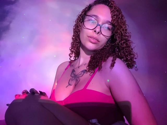 LizzaMonroe-hot with curly brown hair, glasses, nose ring, butterfly tattoo on chest, pink top, sitting, purple and pink lights, and a confident, slightly pouty expression.