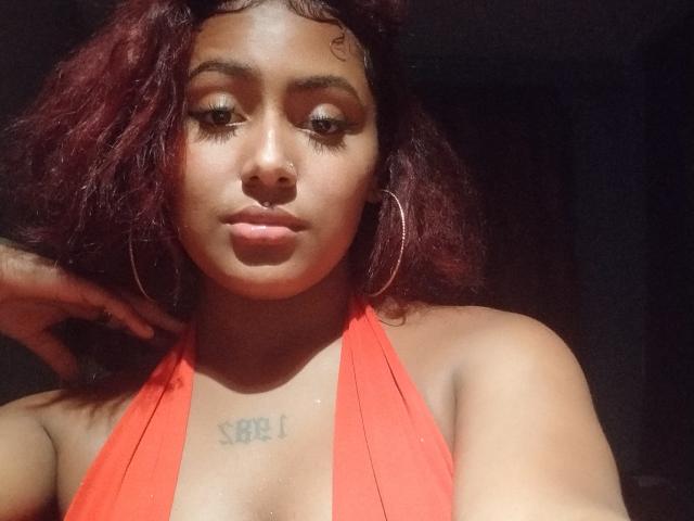 AmberQueeny with dark red curly hair, wearing a red halter top, showing her tits, hoop earrings, and a tattoo under her chest. She has a nose ring and is looking down.