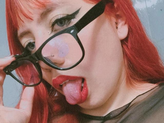 ShelbyShea met rood haar, zwarte bril, neusring en zwarte eyeliner, die haar tong uitsteekt met haar rode lippen, draagt een zwarte top en raakt haar bril aan..