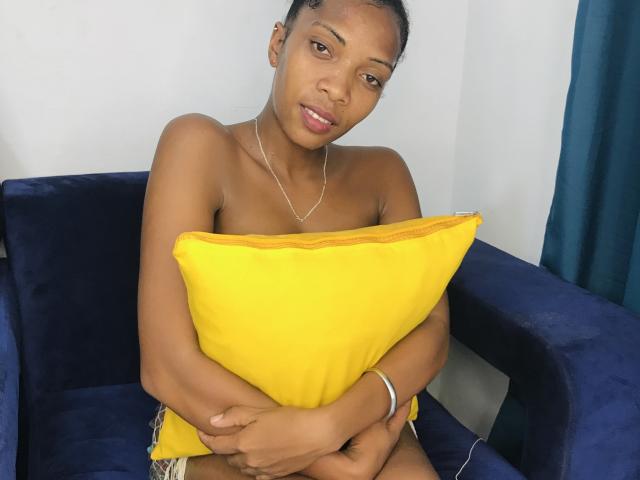 AntonniaMagik with dark skin, small tits, and no clothes, holding a yellow pillow, sitting on a blue chair, looking at the camera with a slight smile.