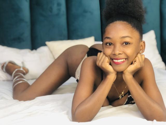 Jaurita with dark brown skin, smiling, lying on a bed, hands under chin, wearing black and white lingerie, gold necklace, curly afro, white bed, blue velvet headboard.