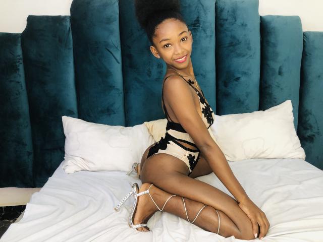 Jaurita with dark skin, big ass, in a black and beige lace-up bodysuit, sitting on a bed, smiling, wearing silver strappy heels, with blue velvet headboard.