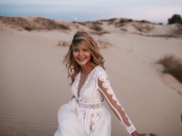 EolantaMarina with blonde wavy hair, smiling, wearing a white lace dress, has tits visible through sheer sleeves, standing in a sandy dune landscape during sunset.