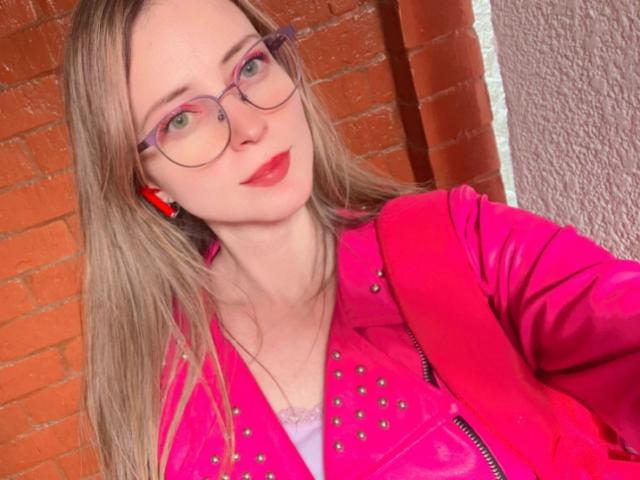 AmbeerWood with pink glasses, red lipstick, and pink leather jacket, showing off her tits in a selfie. She's got long blonde hair and a cute face.