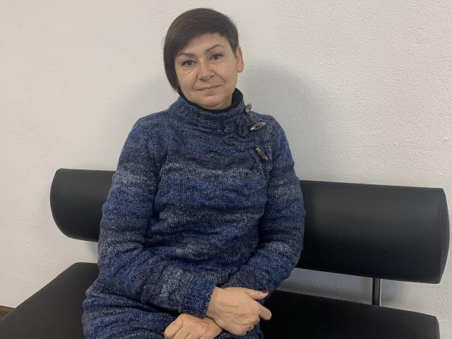 ZorynaYasna, a middle-aged woman with short brown hair, sits on a black cushioned bench. She wears a blue, textured, knitted sweater with button closures on the right side. Her hands are clasped together in front of her. She has a slight, confident smile and is looking directly at the camera. The white wall behind her is plain and unadorned. The composition is simple and focused on ZorynaYasna. The image is clear with natural lighting.