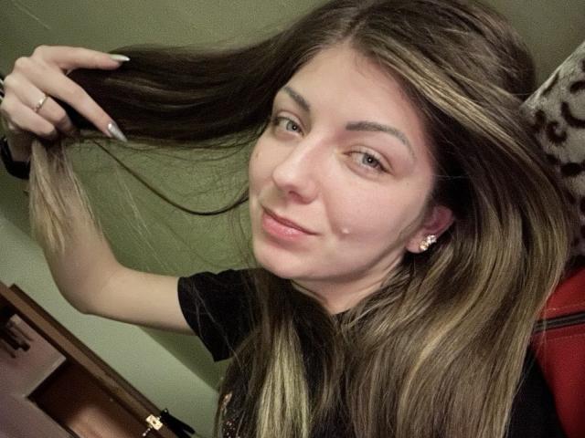 EmilyWilsonsy has long, straight brown hair with blonde highlights, which she holds up with her right hand. She has fair skin, light brown eyes, and a small mole on her right cheek. She wears a black t-shirt and has a subtle, confident smile. Her left hand, with long, pointed nails, is visible. She has small, gold stud earrings. The image has a casual, intimate feel with a warm, greenish background and a red chair in the lower right corner. EmilyWilsonsy's facial features are soft, and her expression is relaxed.