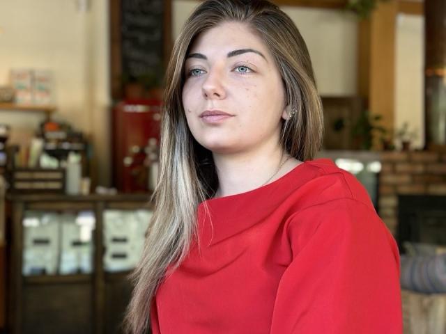 EmilyWilsonsy has fair skin, long straight blonde hair, and green eyes. She wears a red hoodie. Her expression is neutral, with a slight smile. The photo has natural light, highlighting her features. The background is blurred, featuring a mix of wooden and metallic elements, with a chalkboard and shelves. EmilyWilsonsy's makeup is minimal, with subtle eyeliner and light lipstick. The image is clear, focused on EmilyWilsonsy, and has a casual, everyday vibe.