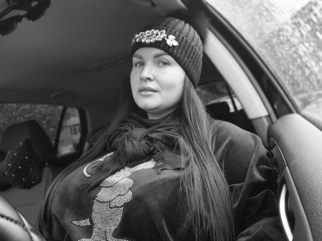 Black and white photo of GeorgiaLerox with large boobs, wearing a knit beanie with flowers, long straight hair, and a dark hoodie with a patterned design, sitting in a car.